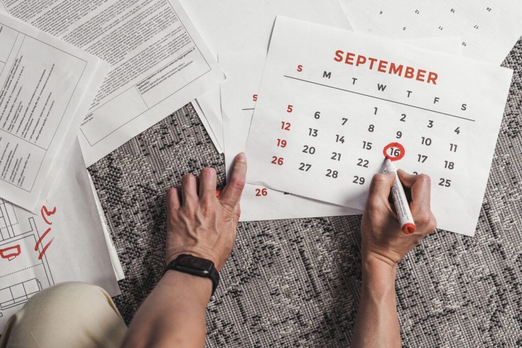 Hands holding a calendar with September dates encircled, surrounded by documents on a carpet.